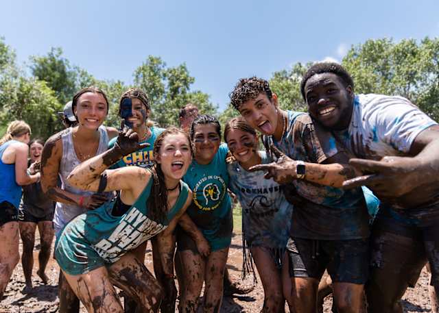 Group of young people smiling and covered in mud during an outdoor team-building activity on a sunny day