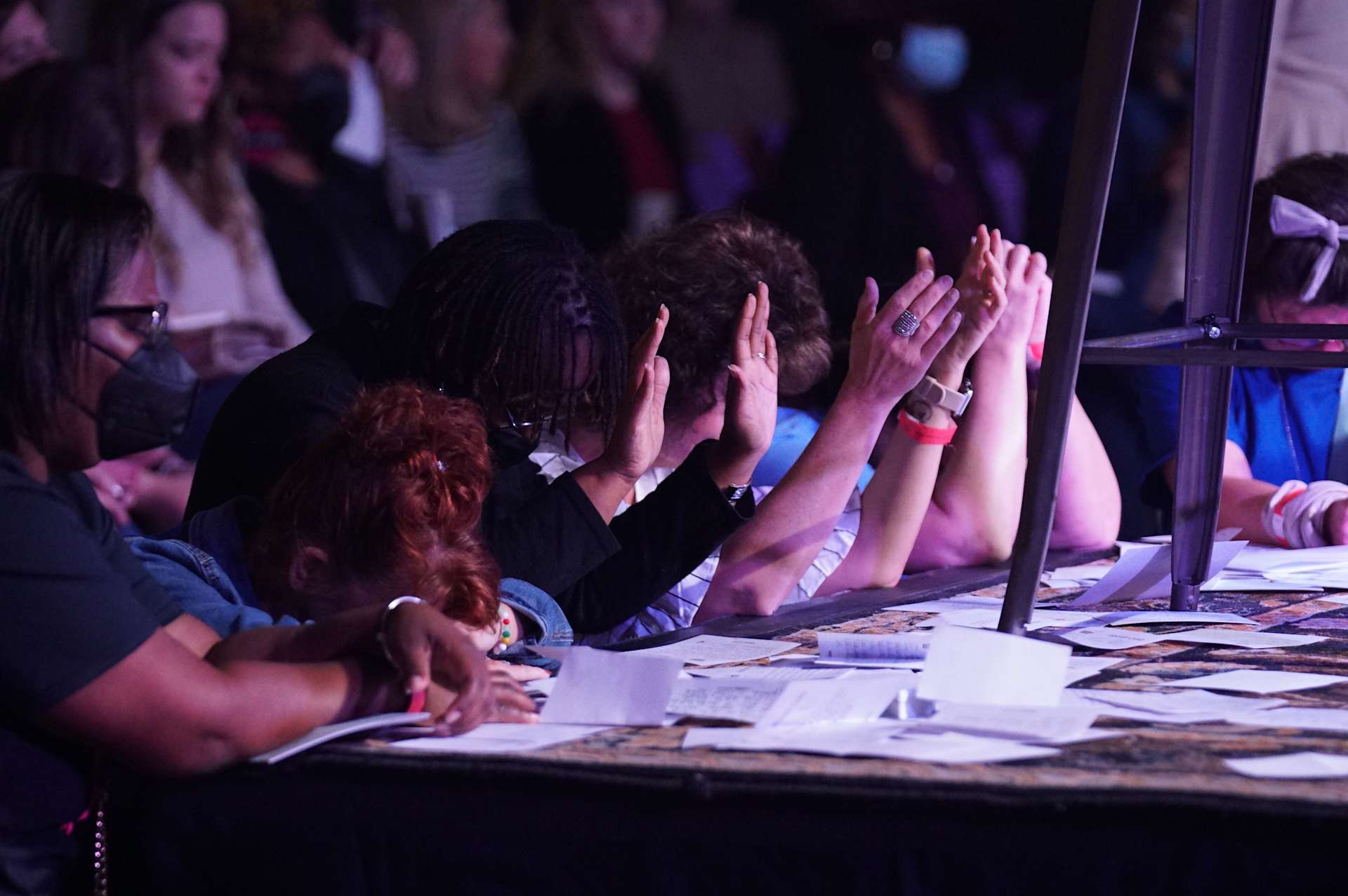 Attendees praying with raised hands, some seated at a table with papers in front of them