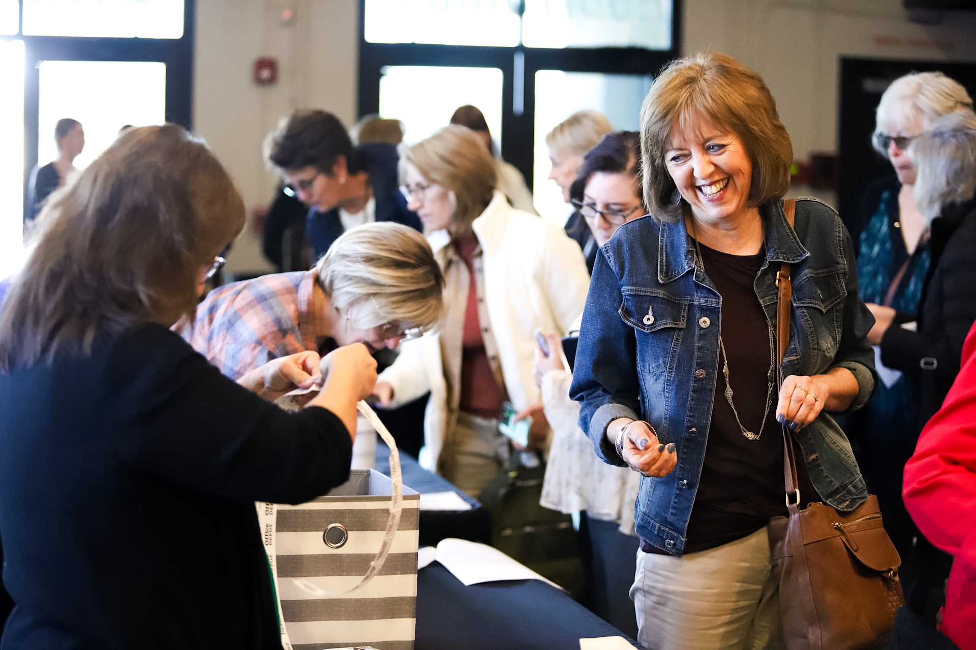 A group of women smile and chat while gathered at an registration table, as others in the background interact