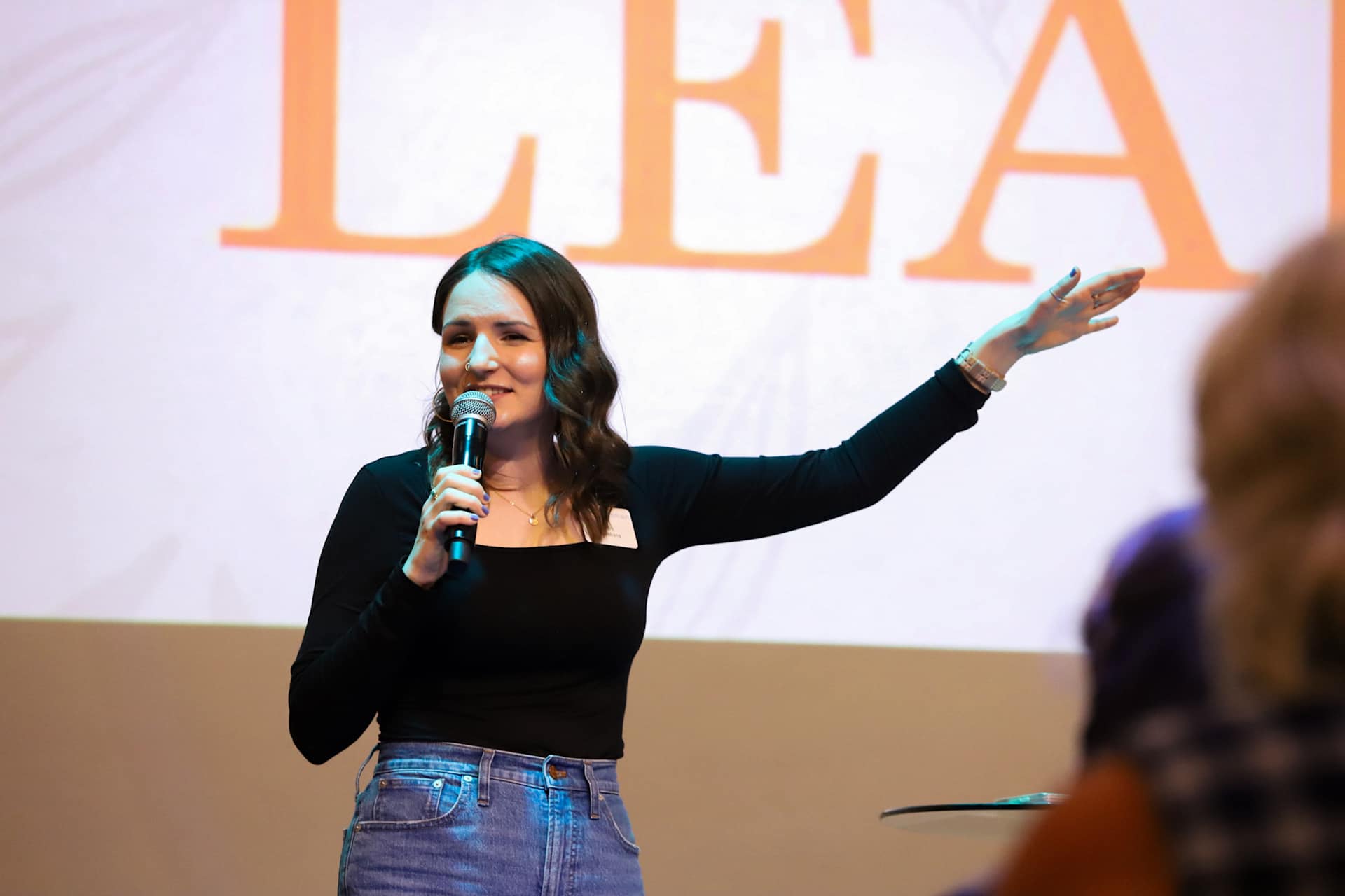 A woman holding a microphone and gesturing while speaking, with the word "LEAD" projected in the background