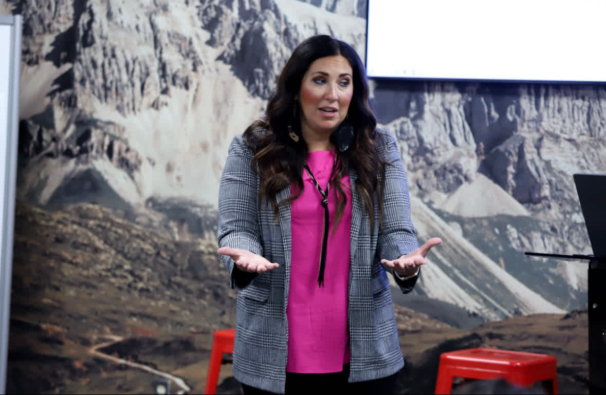 A woman standing in front of a mountain backdrop, gesturing with open hands while speaking