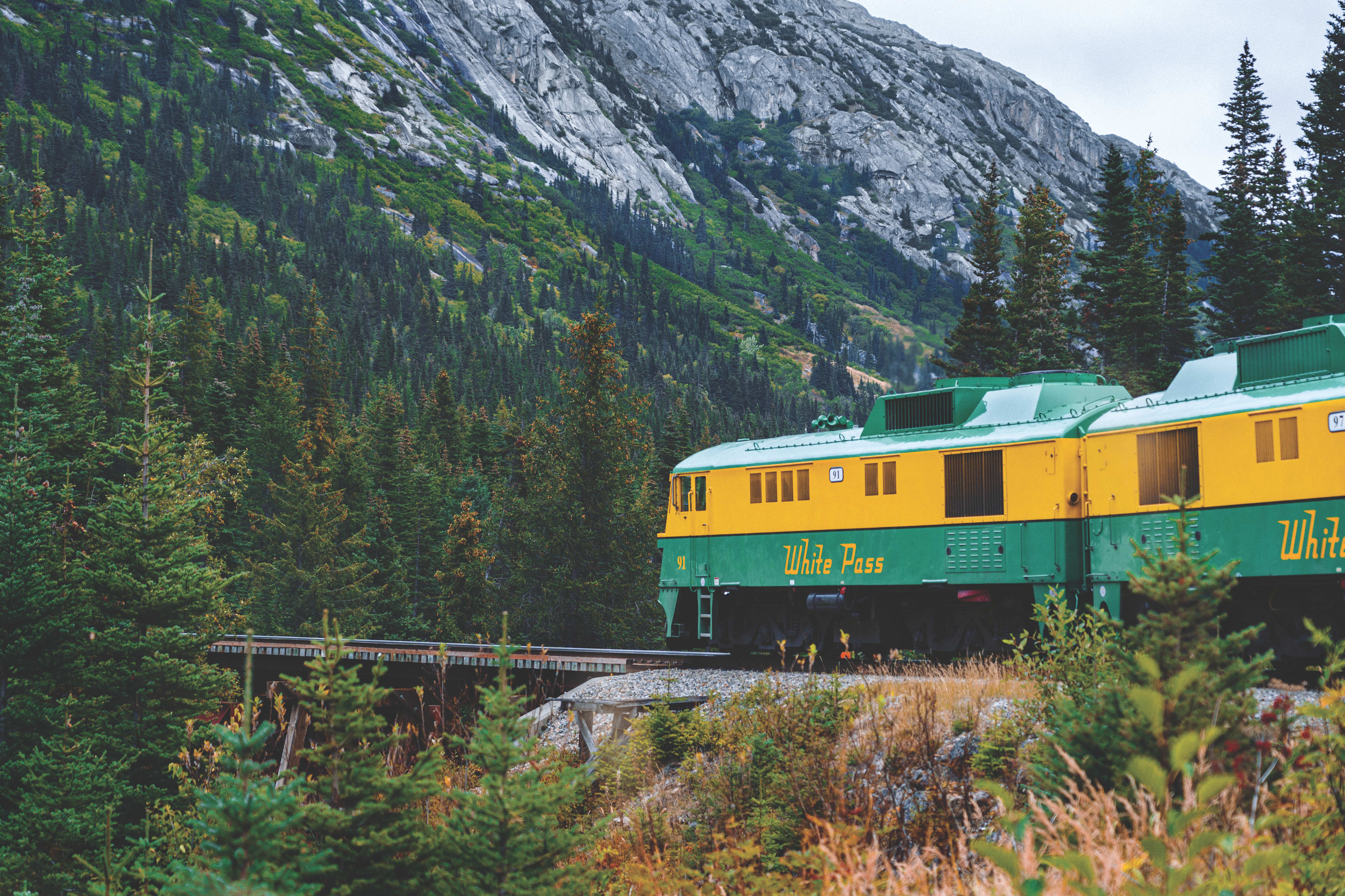 yellow train traveling along mountains with rocks and pine trees