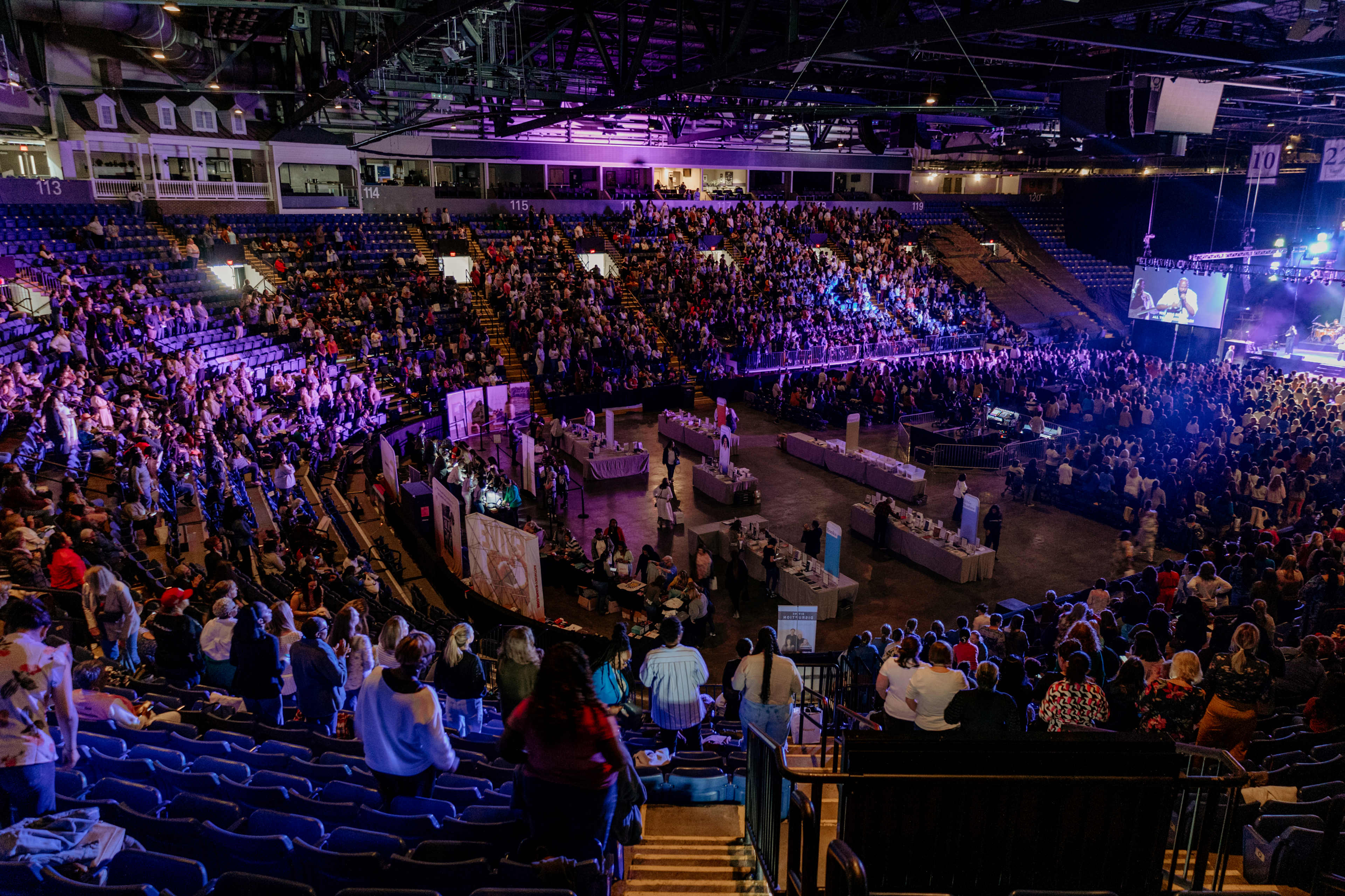 A vibrant crowd fills a large arena for an event, with purple lights illuminating the space