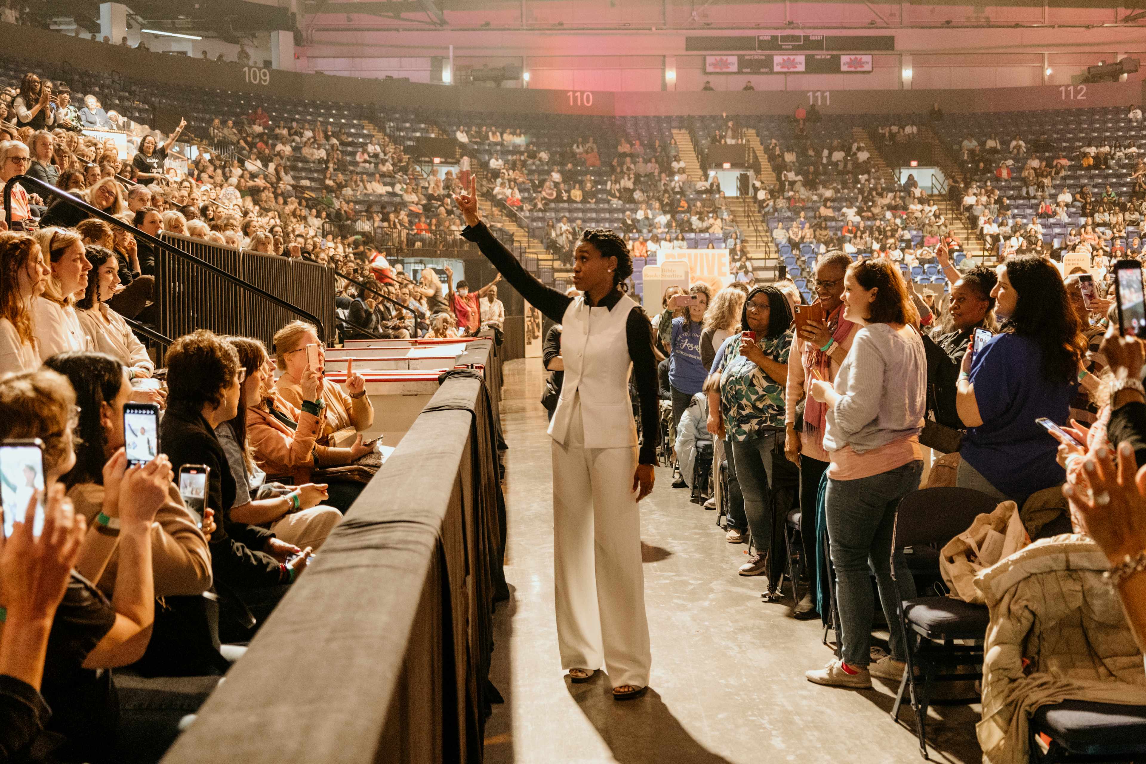 Priscilla Shirer in a white outfit engages with an audience at a large event in an indoor arena