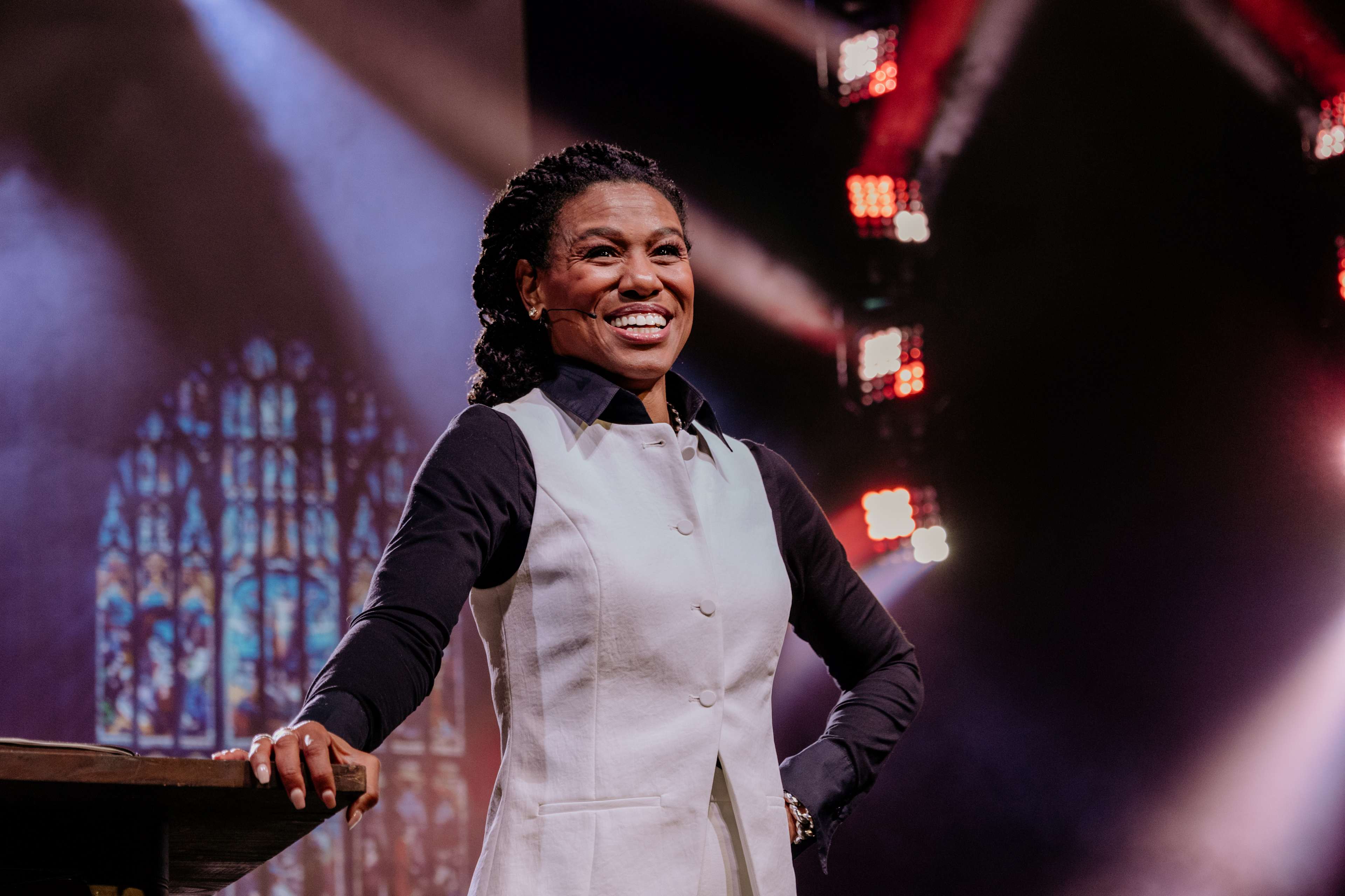 Priscilla Shirer in a light-colored vest smiling confidently on stage, with vibrant lighting creating an uplifting and energetic atmosphere