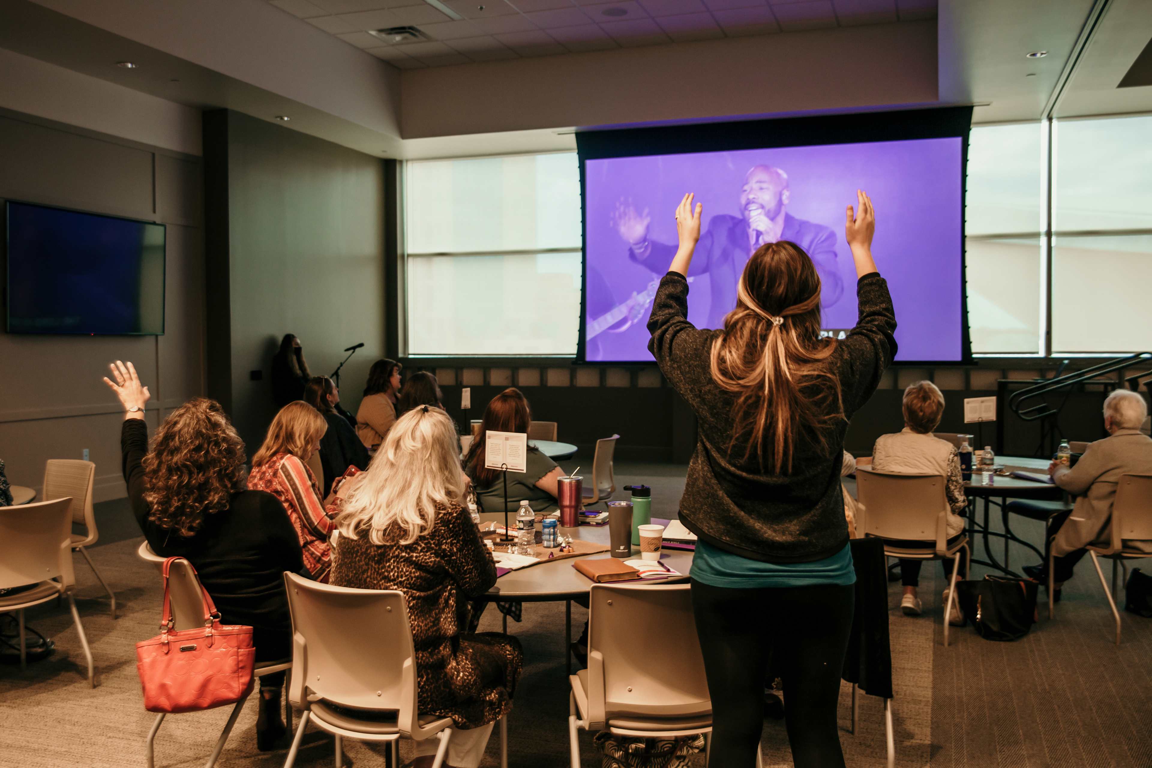 Attendees in a conference room participating in worship while watching a large screen with a man leading worship