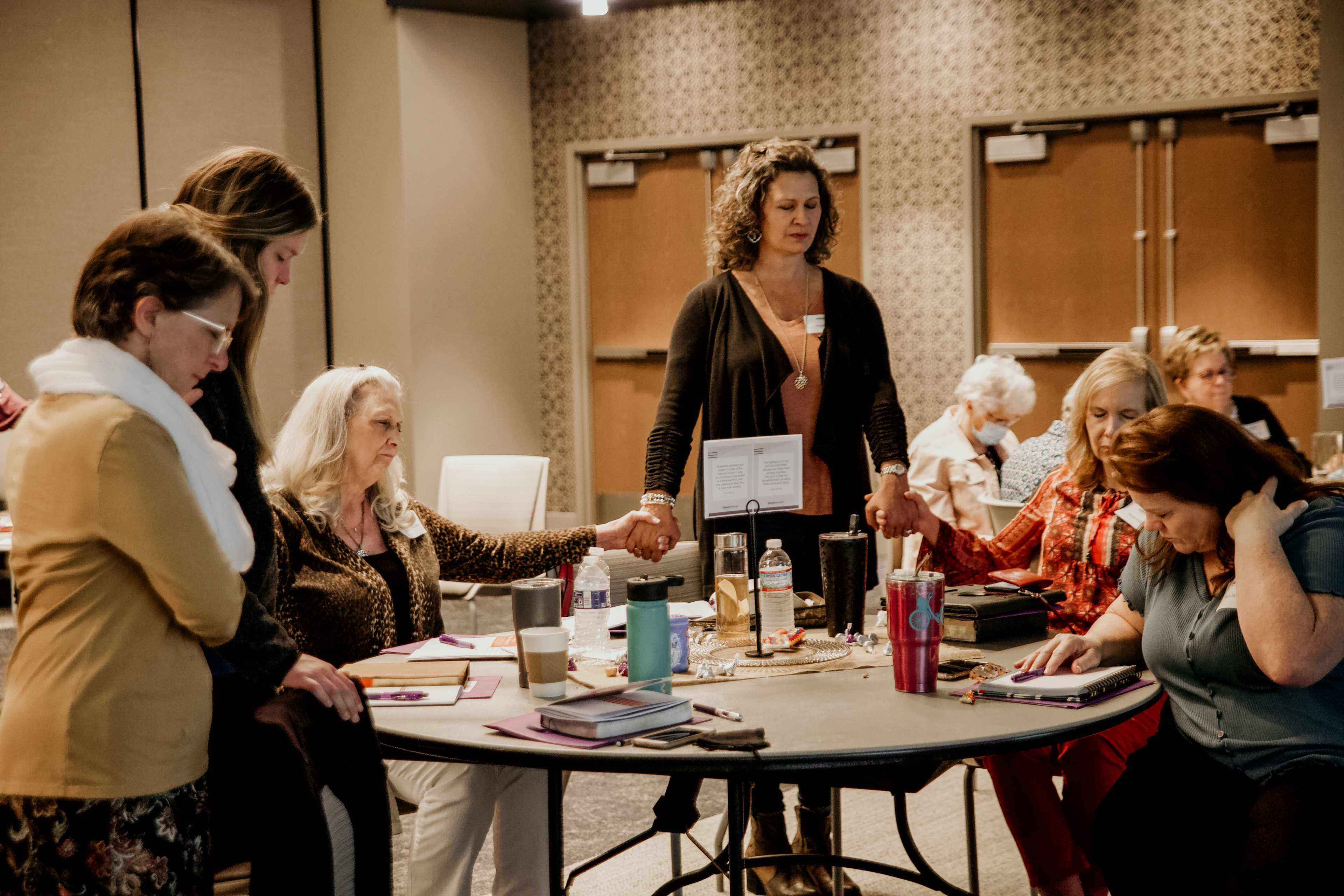 A group of women standing in a circle, holding hands in prayer at a conference table