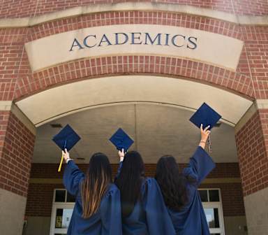 3 women graduates holding their graduation cap under an academics building