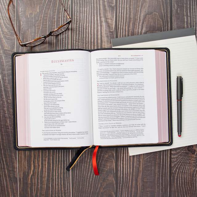 Open Bible on a wooden desk with glasses and a notebook.